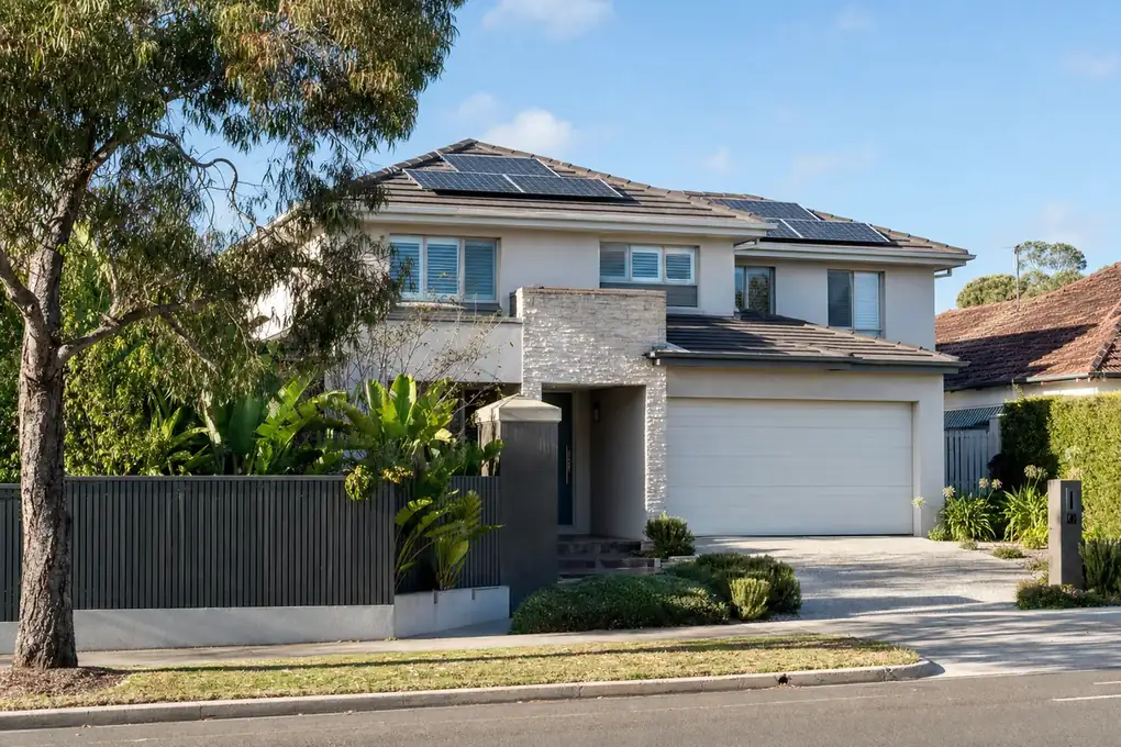 Home battery mounted on the garage wall of a Mentone bayside home, with rooftop solar panels and Port Phillip Bay visible through the coastal tea-trees